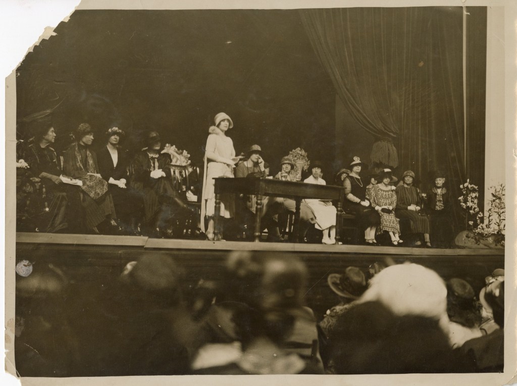 The Duchess of York in a white outfit and hat, standing behind a desk on the conference platform surrounded by other speakers