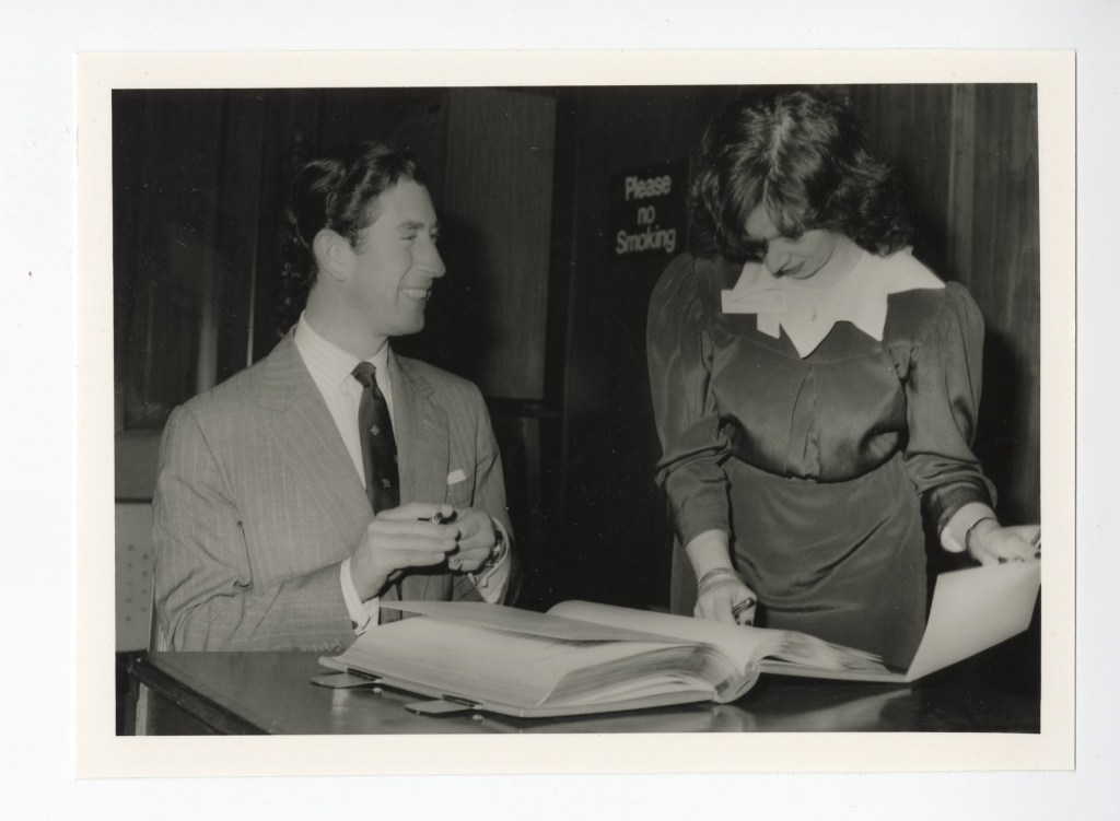 The Prince of Wales sitting at a desk and smiling as he holds a pen to sign a book. The book is being held by a woman who is looking down at the page.